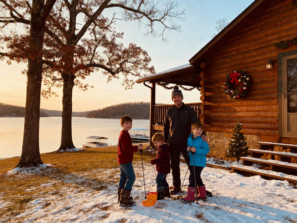 A cozy family enjoying winter activities by a cabin at Holiday Island, with children playing in light snow, Table Rock Lake visible in background, warm evening light, Christmas decorations, Ozark Mountains setting, professional travel photography style