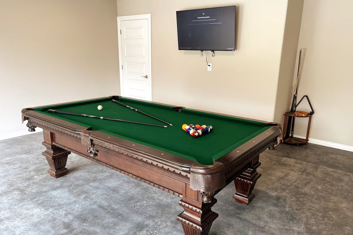 Pool table with green felt and billiard balls in a room with a television mounted on the wall.